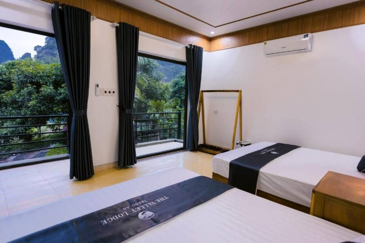 Habitación de hotel con dos camas, un balcón con cortinas y vistas a la vegetación, además de un equipo de aire acondicionado.