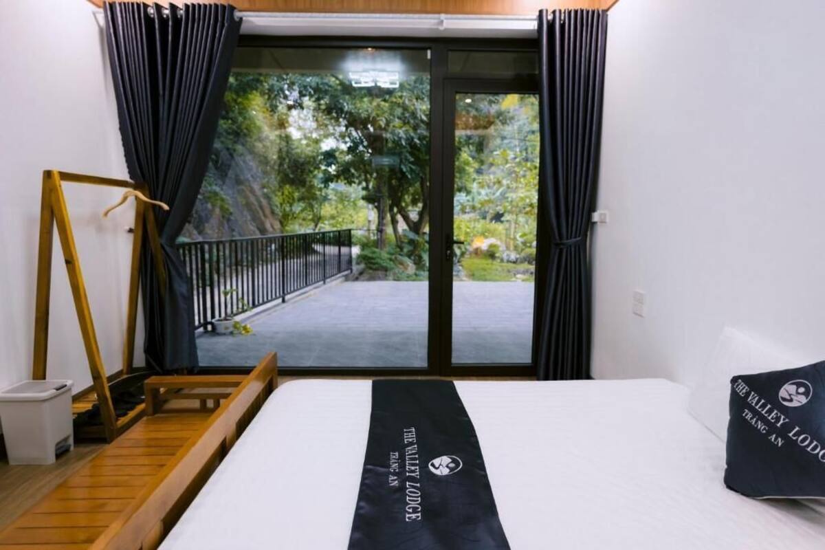 Habitación de hotel con cama, banca de madera, cortina negra y vista a un jardín a través de un ventanal.