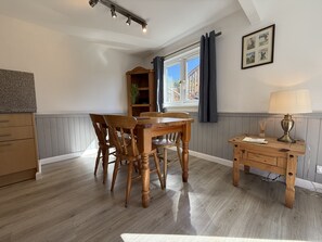 A dining area with a wooden table and chairs, a lamp, a bookshelf, and a window with curtains. - Gigha - a chalet that sleeps 4 guests in 2 bedrooms (Oban)