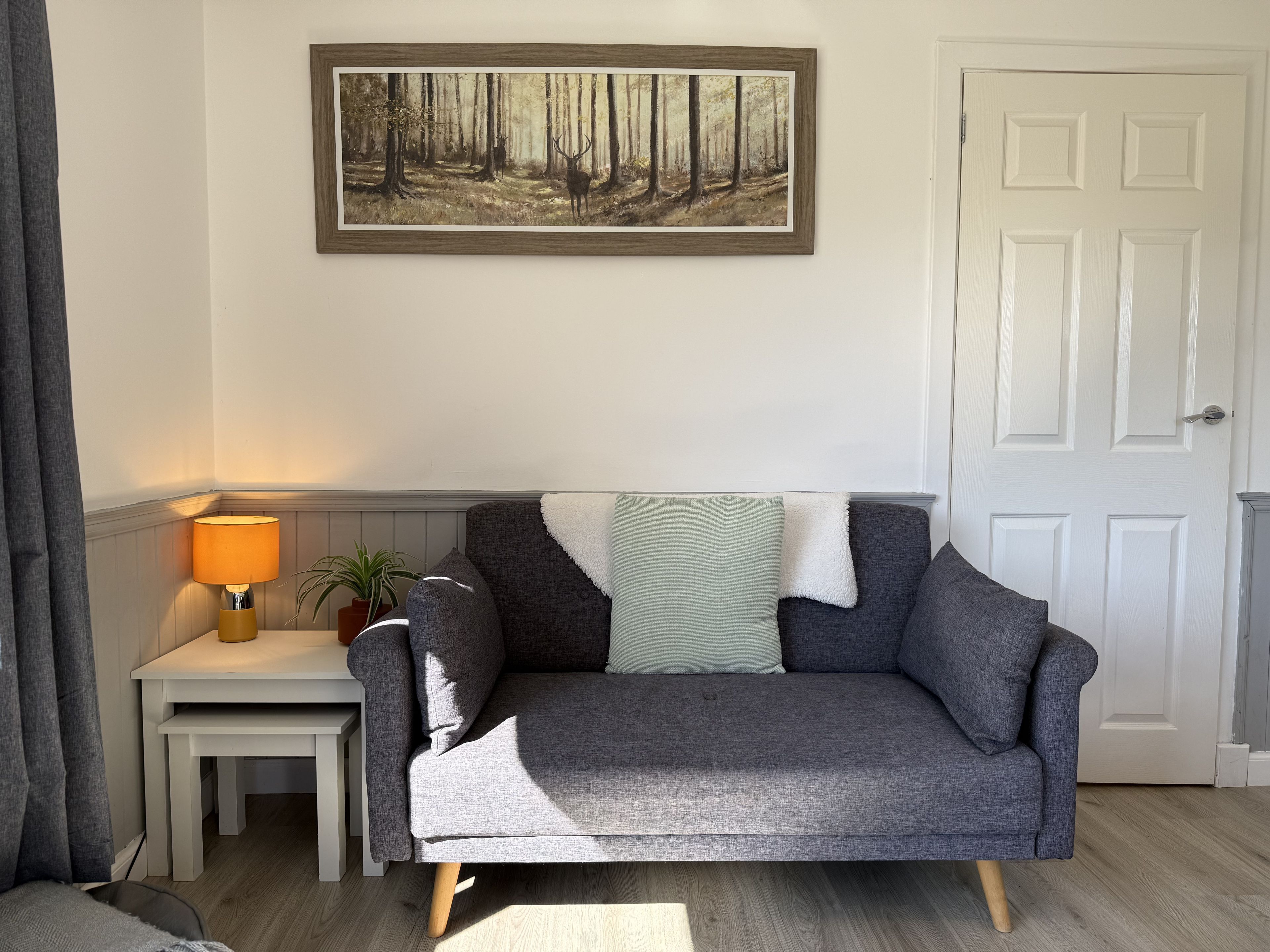 A living room with a grey sofa, a small table with a lamp, a framed painting of a forest, and a white door.