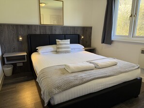Een opgemaakt bed met een gewatteerde deken, een nachtkastje met een lamp, en een raam met gordijnen.