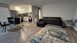 A living room with a black leather sofa, a glass coffee table, a dining area with a glass table and chairs, and a kitchen in the background.