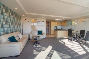 Ein modernes Wohnzimmer mit einer Couch, einem Couchtisch und einem Essbereich mit Stühlen. Im Hintergrund ist eine Küche zu sehen.