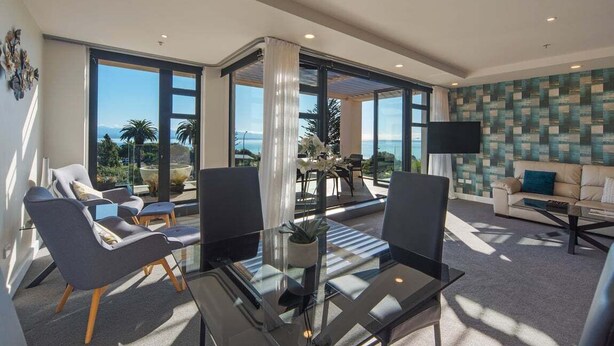Un salon moderne avec une table en verre, deux fauteuils, un canapé, un téléviseur et de grandes fenêtres donnant sur la mer.