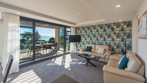 Un salon moderne avec un canapé, une table basse et un téléviseur. Une grande fenêtre offre une vue sur l’extérieur.
