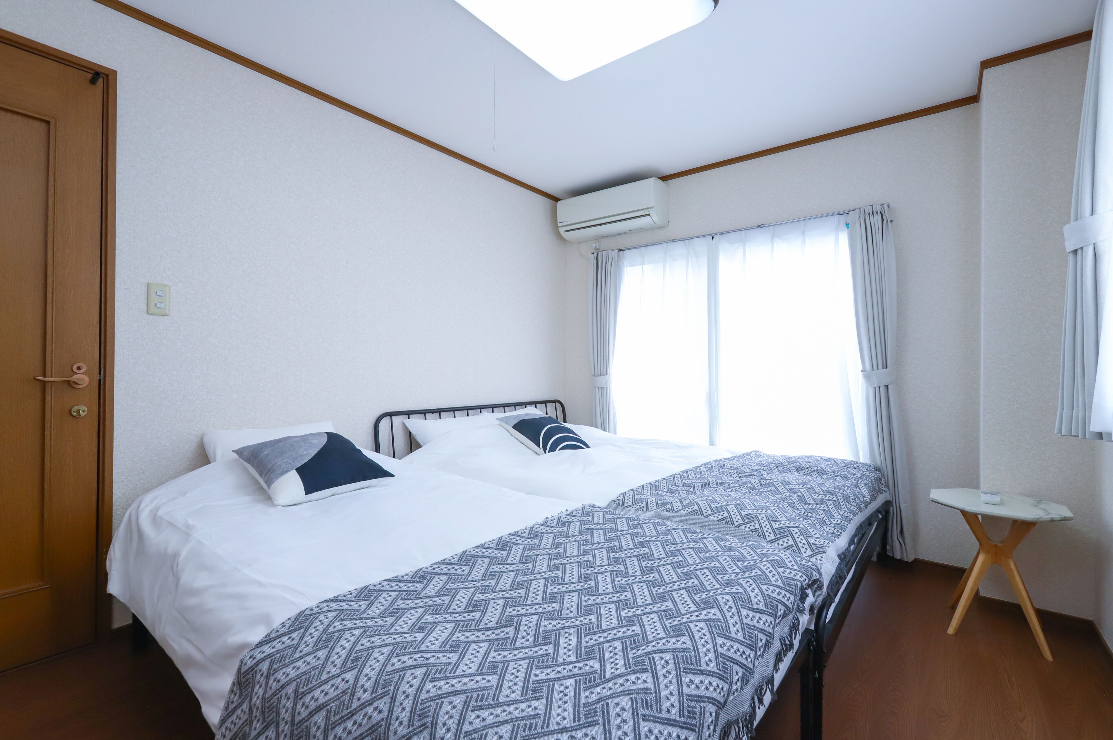 A hotel room with a bed, a small table, an air conditioning unit, a window with curtains, and a wooden door.