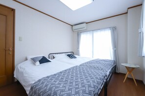 A hotel room with a bed, a small table, an air conditioning unit, a window with curtains, and a wooden door. - 3-Bedroom House, Near Ikebukuro and a Shopping Arcade (Nukui, Nerima City)