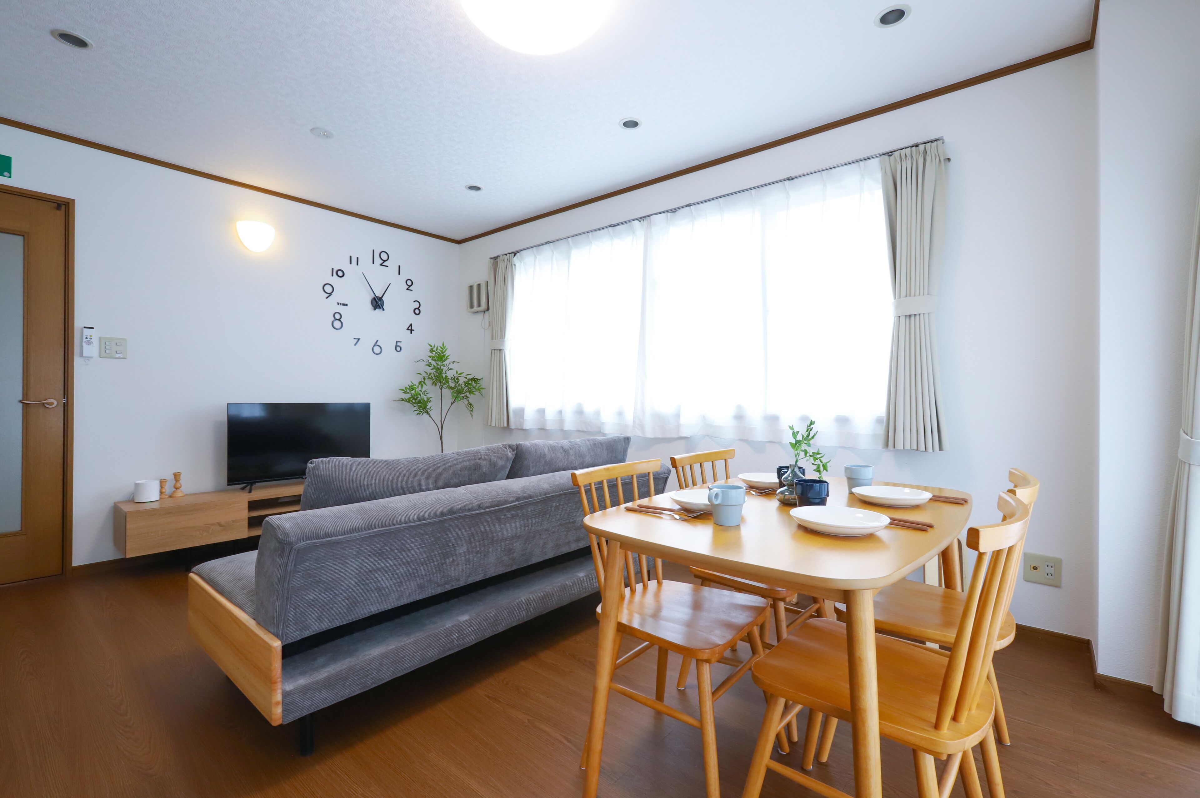 A modern living room with a sofa, dining table, and chairs. There is a flat-screen TV on a stand, a clock on the wall, and a window with curtains.