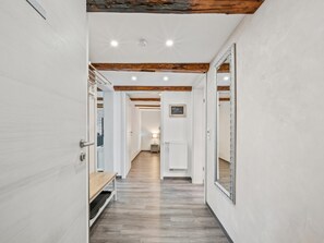 Ein Flur mit Holzboden, weißen Wänden und freiliegenden Holzbalken. Eine Bank, ein Heizkörper und ein gerahmter Spiegel sind an der Wand angebracht.