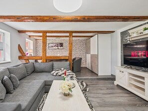 Ein modernes Wohnzimmer mit einem grauen Sofa, einem Couchtisch mit Blumen, einem Fernseher, der 'NETFLIX' anzeigt, und freiliegenden Holzbalken an der Decke.