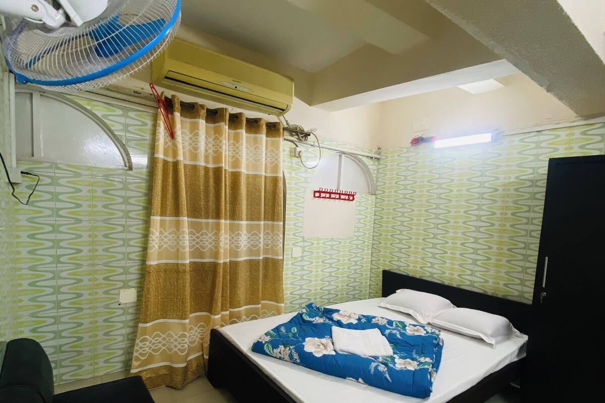 A room with a bed, a desk, a chair, a standing fan, an air conditioning unit, and patterned walls.