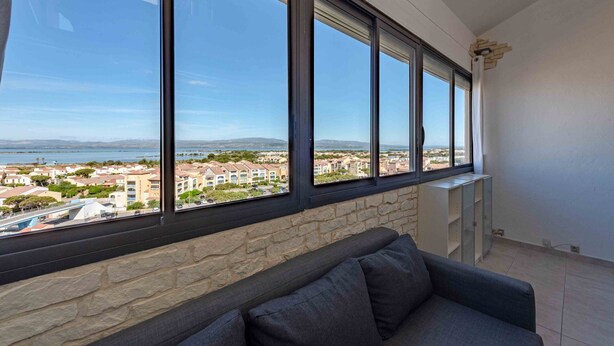 Quarto com uma grande janela com vista para a costa, uma parede de pedra e um sofá cinzento.