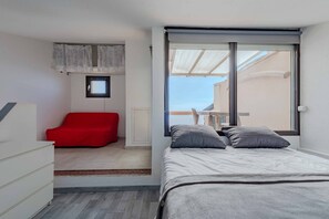 A bedroom with a bed, a red chair, a small window, and a view of the outside.