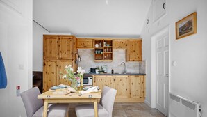 A compact kitchen with wooden cabinets, a dining table with two chairs, and a small island with a sink. - The Granary - Swimming pool, Parking, Garden, BBQ (Georgeham)