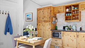 A kitchen with wooden cabinets, a microwave, and a dining area with a table and chairs. - The Granary - Swimming pool, Parking, Garden, BBQ (Georgeham)