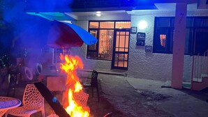 A fire pit with burning logs in a courtyard, surrounded by outdoor furniture and a building with a door and windows in the background. - Pankaj Homestay (Shimla)