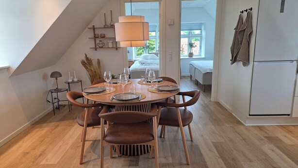 A dining area with a round table set for six, wooden chairs, a lamp, and a view of a bedroom through an open door. - Bright modern holiday apartment on Samsø (Samsø)