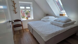 A bedroom with a sloped ceiling, a bed with white bedding, a wooden chair, a window with a view of buildings, and a small table with a basket. - Bright modern holiday apartment on Samsø (Samsø)