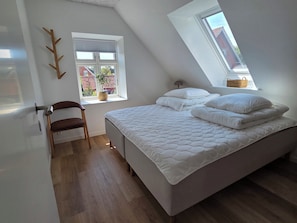 A bedroom with a sloped ceiling, a bed with white bedding, a wooden chair, a window with a view of buildings, and a small table with a basket.