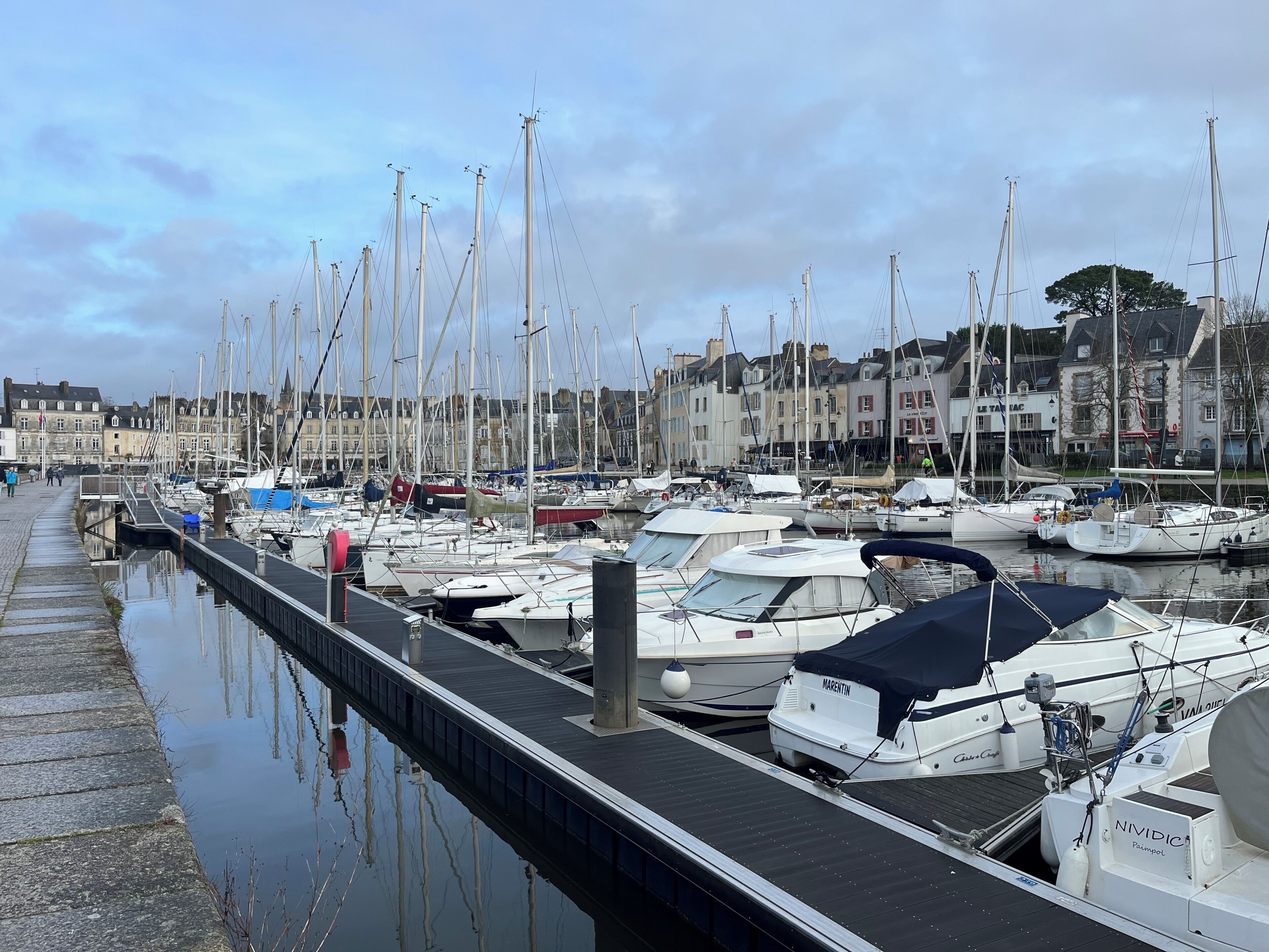 A marina with boats docked, a wooden walkway, and buildings in the background.