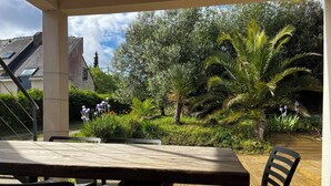 Een terras buiten met een houten tafel en stoelen, omgeven door weelderige begroeiing en bloeiende planten.