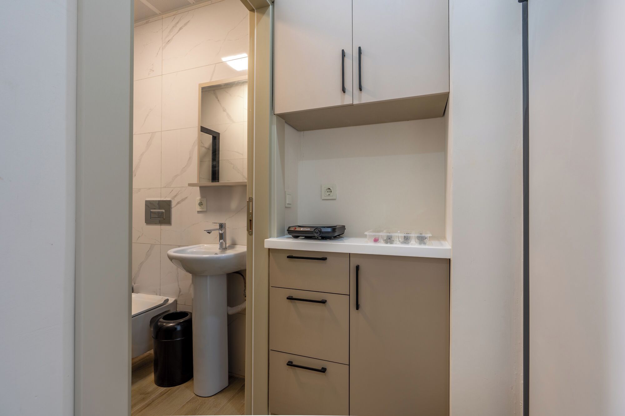 A compact bathroom with a pedestal sink, a trash bin, and a cabinet with a mirror above it.