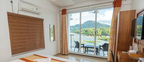 A hotel room with a bed, a TV, a desk, and a view of a river and mountains.