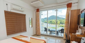 A hotel room with a bed, a TV, a desk, and a view of a river and mountains. - The Anchorage Athirappilly Resort (Chalakudy)