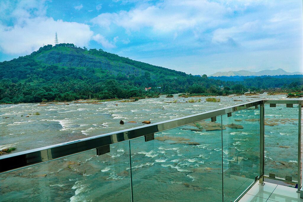 A glass bridge over a river with a view of a green hill and a distant mountain range.