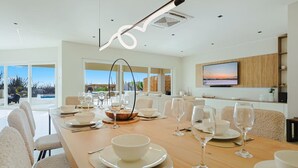 A modern dining area with a long table set for a meal, a television mounted on the wall, and a view of the outdoors. - LUX 4BR Modern Villa • Pool Oasis • Villa Graziano (Noord)