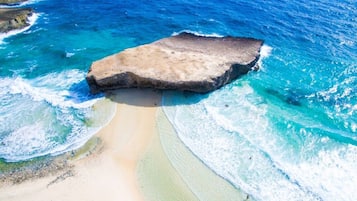Une plage avec une grande formation rocheuse, une eau turquoise cristalline et un rivage sablonneux.