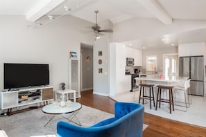 Ein modernes Wohnzimmer mit Flachbildfernseher, einem blauen Sessel, einem kleinen weißen Tisch und einer Küchenzeile mit weißen Schränken und Edelstahlgeräten.
