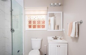 Ein Badezimmer mit Duschkabine aus Glas, Toilette, Waschbecken, Spiegel und Fenster mit ornamentiertem Sichtschutz.