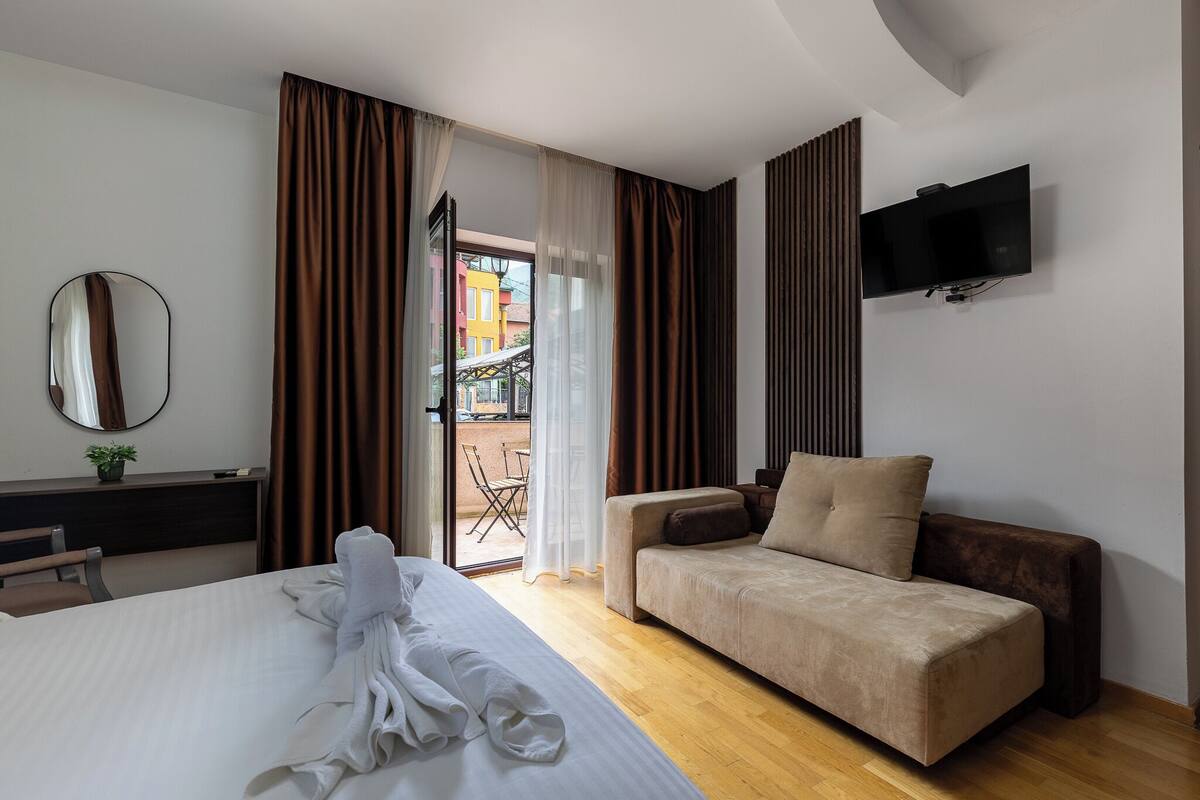 A hotel room with a bed, a sofa, a TV, a desk with a plant, and a view through a door.