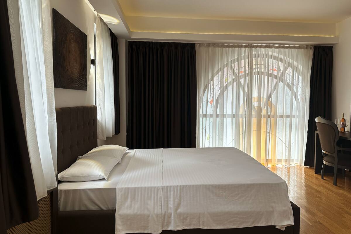 A hotel room with a large bed, a desk with a chair, and a view through sheer curtains.
