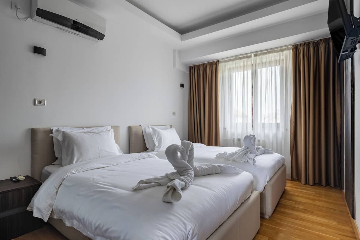 A hotel room with two beds, a flat-screen TV, an air conditioning unit, and a window with curtains.
