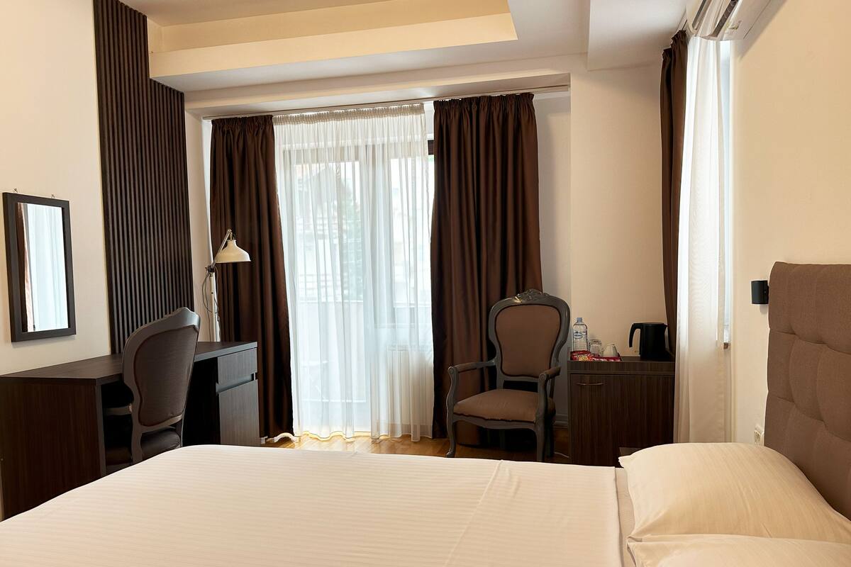 A hotel room with a bed, a desk, a chair, a lamp, and a window with curtains.