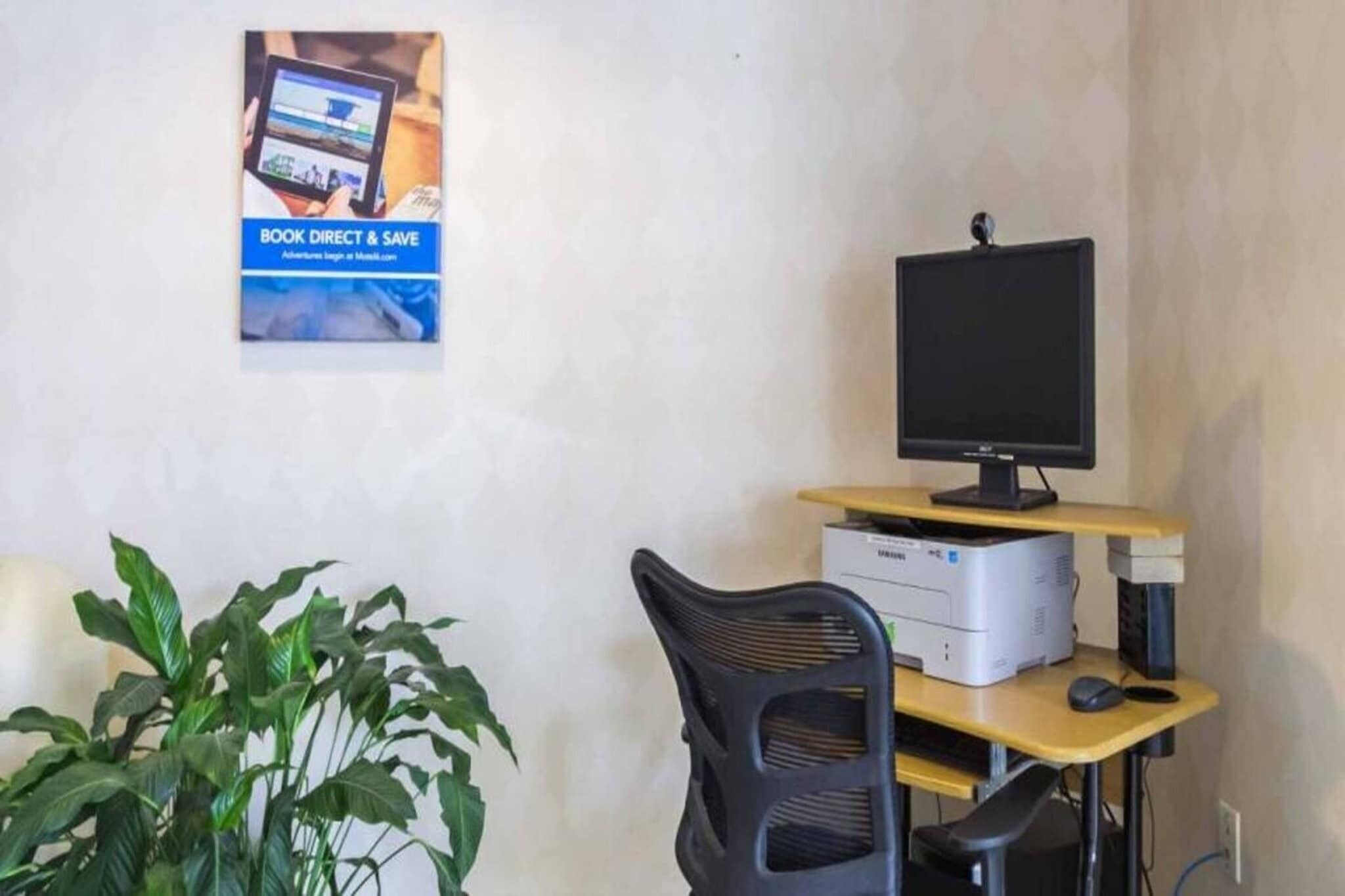 Ein Schreibtisch mit Computermonitor, Drucker und Tastatur. Vor dem Schreibtisch steht ein Stuhl. Eine Topfpflanze ist auf der linken Seite zu sehen.
