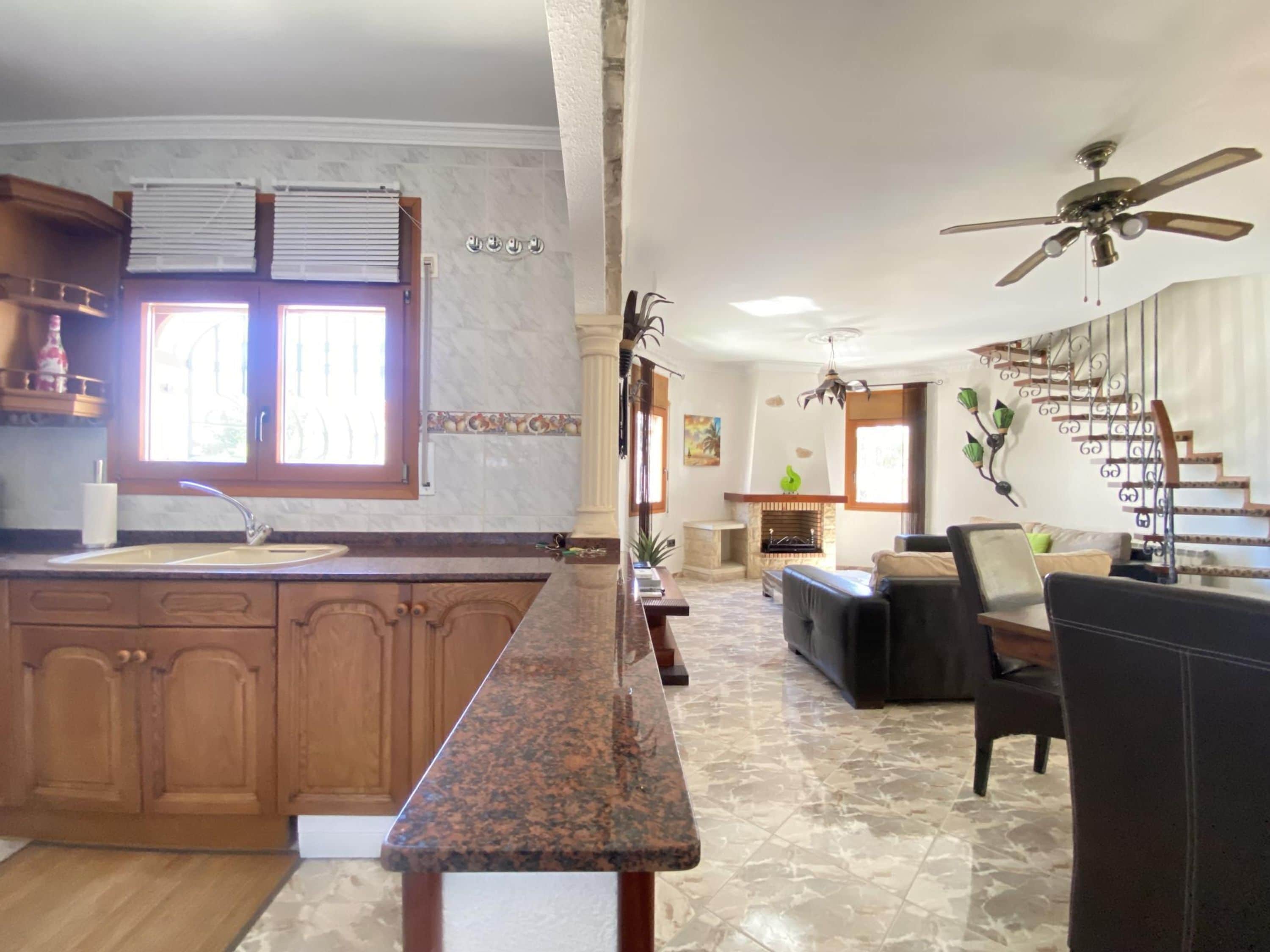 Una cocina con armarios de madera, encimera de granito, zona de comedor con mesa y sillas, chimenea y escalera con barandilla de metal.