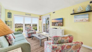 A living room with a sofa, armchair, and coffee table, a view of the outdoors, and decorative items like a sailboat and a lighthouse. - Oceanfront 3BD/2BA-First Floor-Beautifully Decorated-Family Friendly (North Myrtle Beach)
