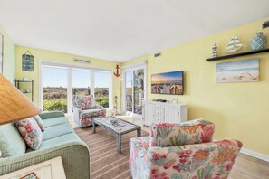 Ein Wohnzimmer mit Sofa, Sessel und Couchtisch, Blick ins Freie sowie dekorative Elemente wie ein Segelboot und ein Leuchtturm.