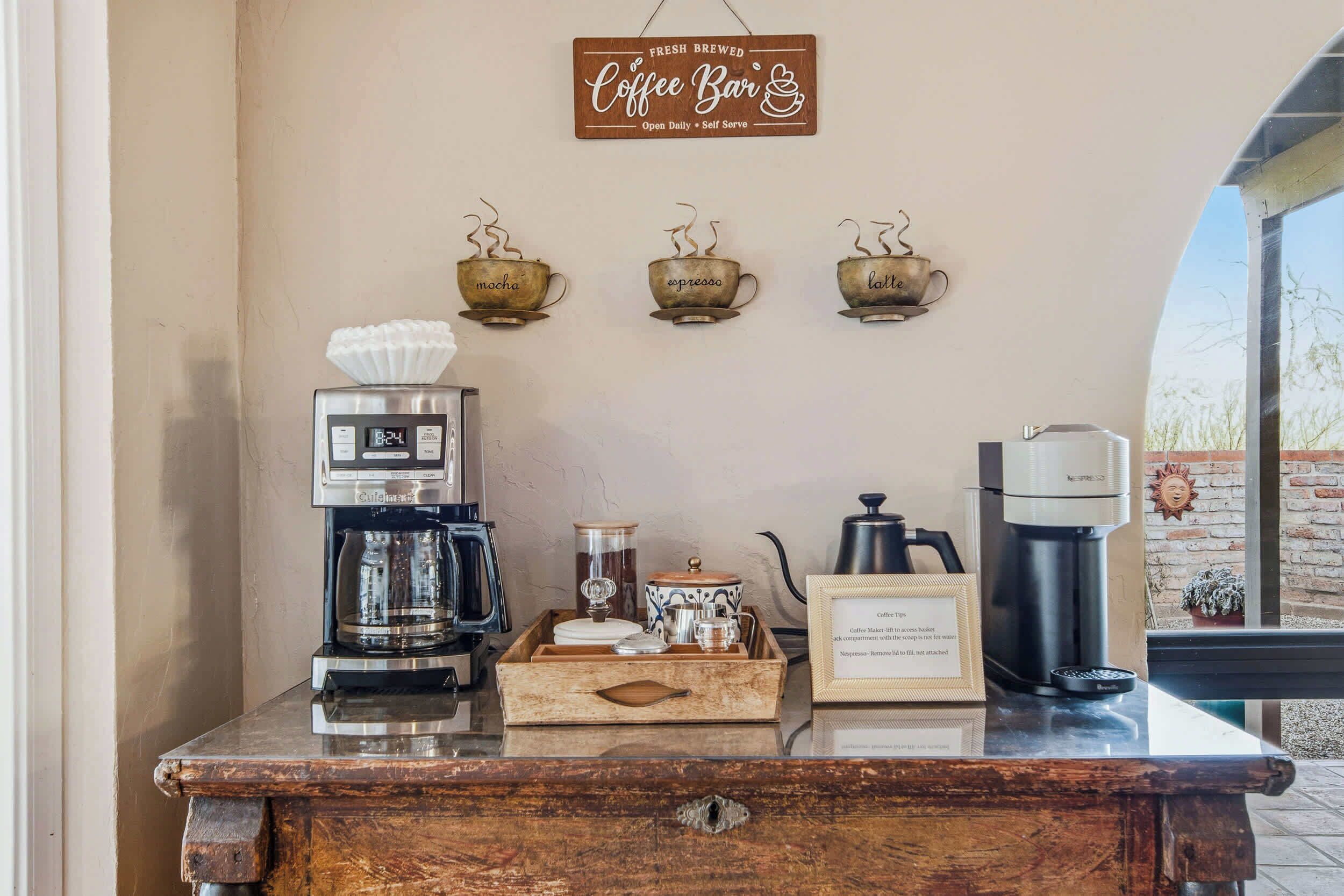 A coffee bar with a sign, three coffee cups, a coffee maker, a milk frother, a coffee grinder, and a tray with coffee cups and a sign.