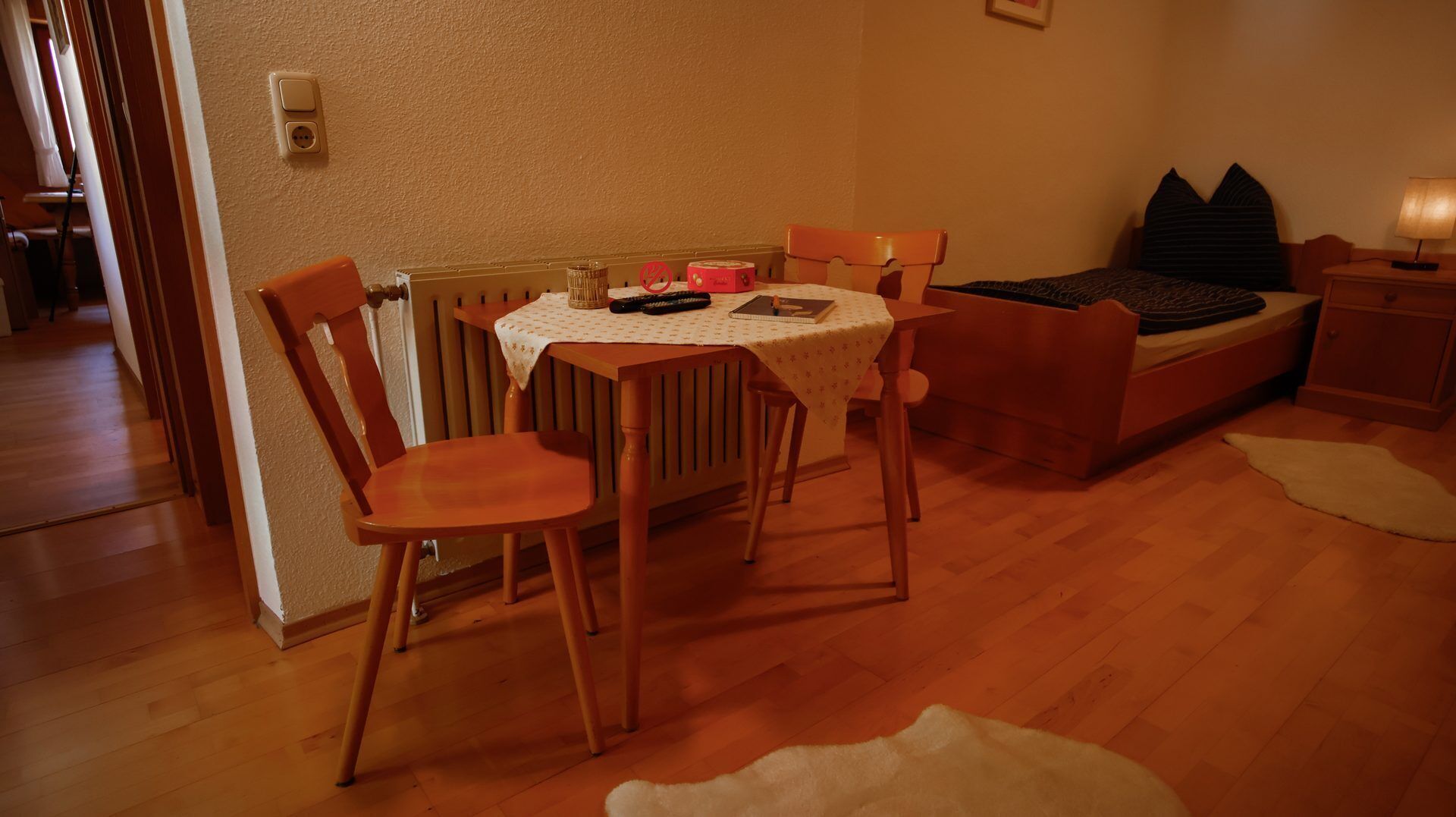 Una habitación acogedora con una mesa redonda, dos sillas de madera, una cama con cabecera oscura, una mesita de noche con lámpara y un radiador en la pared.