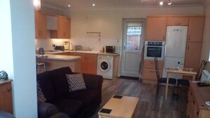 Een ruimtelijk ingerichte leefruimte met een bank, een tafeltje, een wasmachine, een koelkast en een kitchenette.
