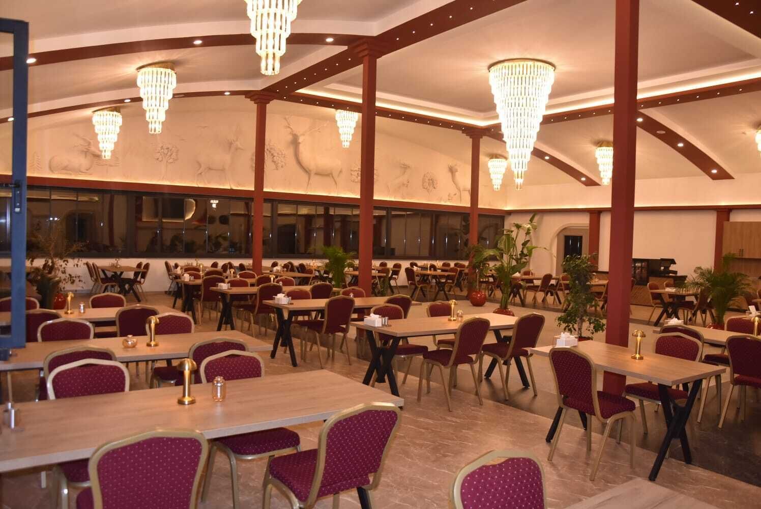 Un restaurant spacieux, doté de nombreuses tables et chaises, de grands lustres et offrant une vue sur la cuisine à travers les fenêtres.