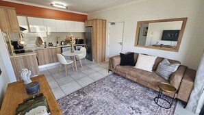 Een moderne keuken met witte kasten, een zithoek met een witte tafel en stoelen, een bruine bank met kussens, en een vloerkleed met een patroon.