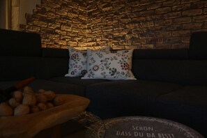 Una sala acogedora con pared de ladrillo, sofá oscuro, cojines estampados y una mesita de madera con un cuenco de nueces.
