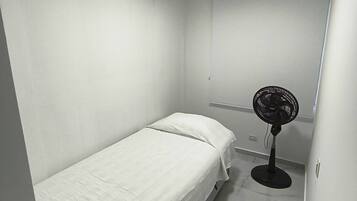 Een kleine kamer met een enkel bed, een witte spreidek, een ventilator en een plafondlamp.