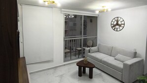 En moderne stue med en hvid sofa, et lille træbord, et dekorativt ur og et stort vindue med udsigt til en balkon.