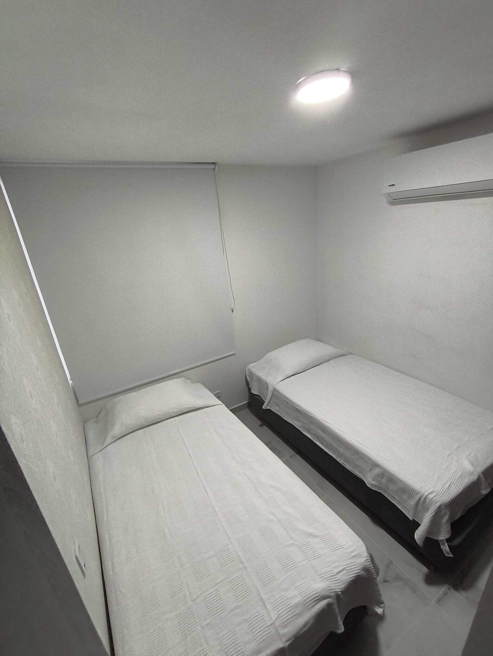 A small, single-room with two single beds, a white ceiling, and a small shelf.
